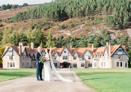 bride and groom standing in front of large country house with Scottish hills surrounding them